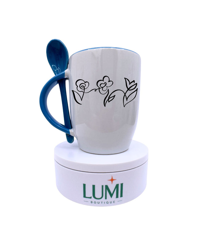 White mug with blue handle and floral design, accompanied by a blue spoon, on a white background with LUMI Boutique branding.