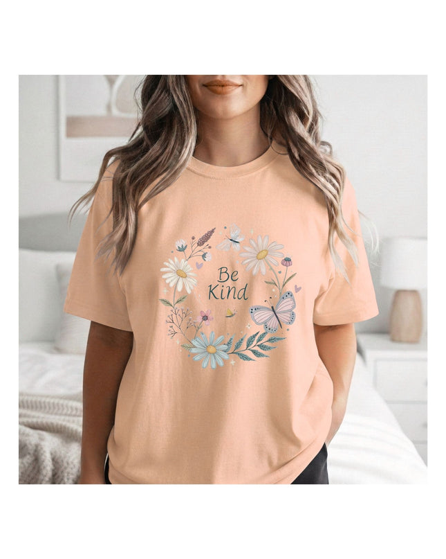 Person wearing a peach t-shirt with floral design and 'Be Kind' text in a bedroom setting